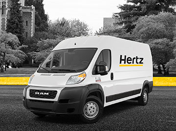 White cargo van with Hertz logo on side and yellow line with trees in the background.