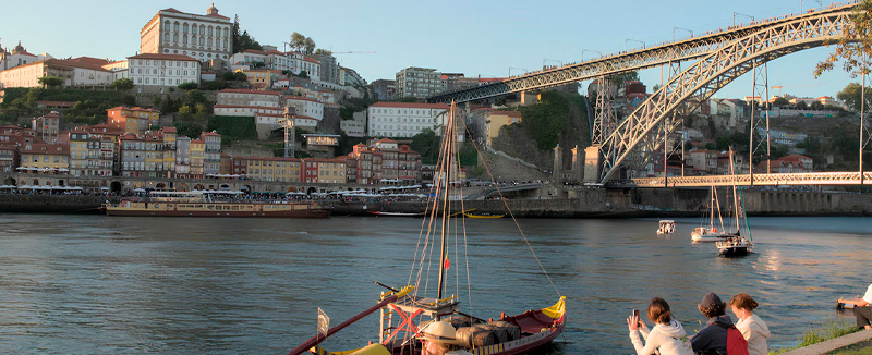 Dom Luís I Bridge in Porto, Portugal, over the Douro River with boats and people enjoying the riverside.