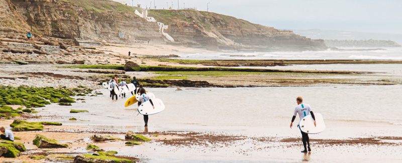 grupo de jovens vestidos com fatos de mergulho e com a prancha de surf na mão a caminhar numa praia com diversas rochas 