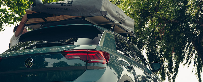 Carro de aluguer Hertz Volkswagen T-Roc verde com tenda de tejadilho montada, estacionado junto a árvores, pronto para uma viagem de aventura em Portugal.