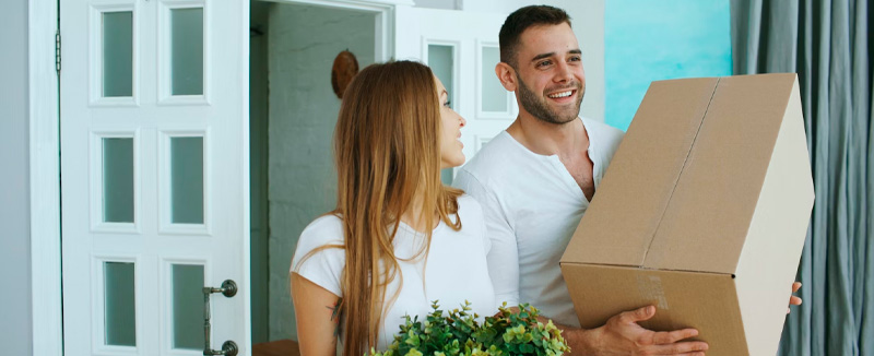 A woman wearing a white T-shirt and pants is setting a cardboard box down on a wooden bench in a room filled with boxes