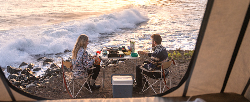 view from inside a tent above the car of a woman and a man sitting on chairs between them at a square table with food and the sea in the background 