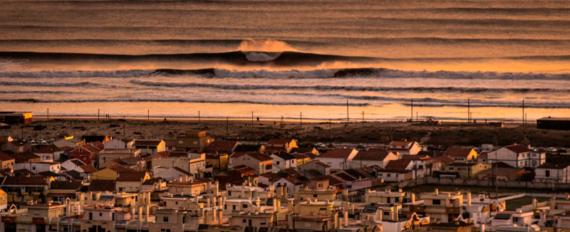 A view of the ocean with waves and houses along the Caparica coast at sunset