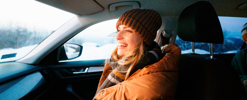 mulher sorridente vestida com gorro, casaco e cachecol dentro de um carro no lugar de pendura e fora do carro neve
