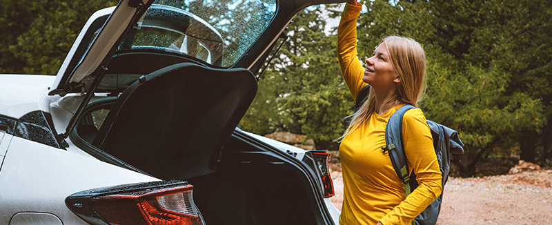 mulher com mochila às costas e camisola amarela está com o braço direito esticado para fechar o porta bagagens de um suv branco