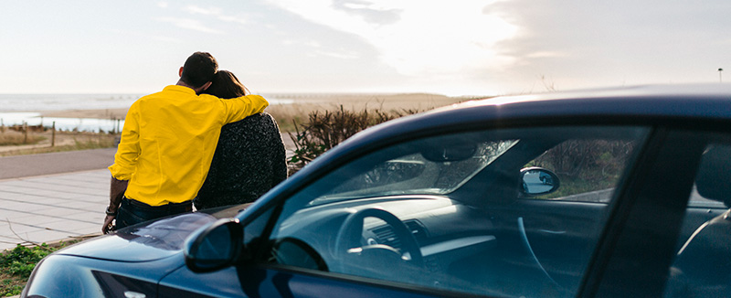 casal virado para uma paisagem desafogada onde o homem que está vestido com um casaco amarelo tem o braço direito envoltada mulher e os dois estão encostado ao carro