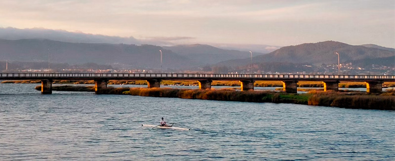 A bridge over a river with a person kayaking in the late afternoon, with mountains in the background