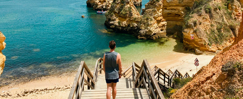 Homem com calções e manga cava de costas a andar na escadaria de madeira da praia do camilo no algarve, na paisagem a areia e o mar cristalino da praia