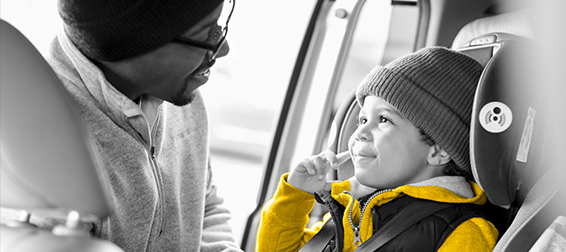 A father wearing a black beanie and glasses smiles as he buckles his young toddler son into a rental car seat.