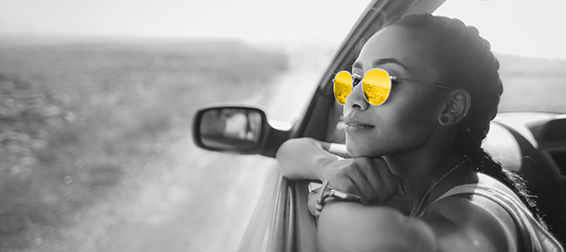 A woman in her 20s wearing round sunglasses rests her chin on her hands as she looks out of a car driver's side window.