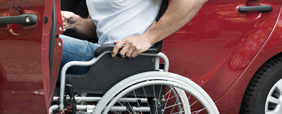 A man wearing a light gray shirt and blue jeans prepares to push himself out of his wheelchair and into a red car.